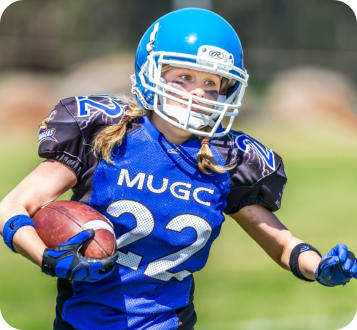 Closeup of a school-aged girl playing American football. She is wearing a blue uniform and helment, and is running with the football tucked under one arm.