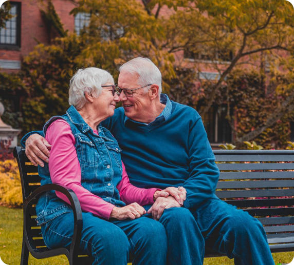 A senior couple sits together outside on a bench on a nice autumn day. The man has his arm around the woman and they looking at each other and smiling.